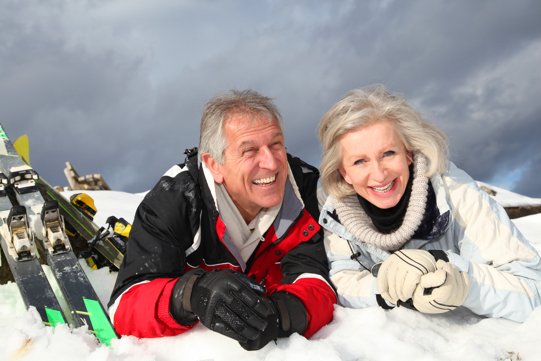 Senioren-Ehepaar liegt mit abgeschnallten Skiern lachend im Schnee.