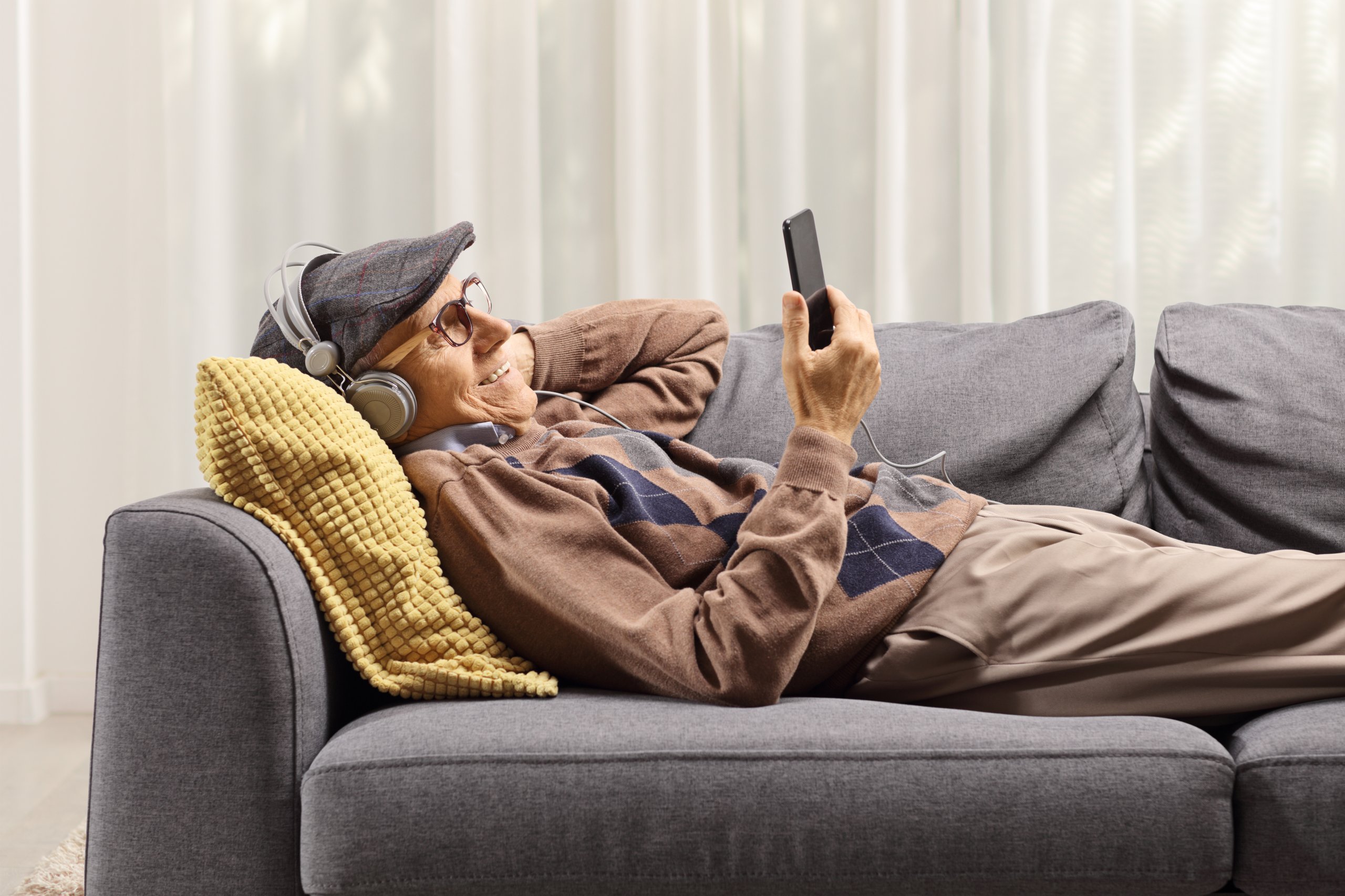 Senior liegt auf Sofa mit Kopfhörern und Smartphone.
