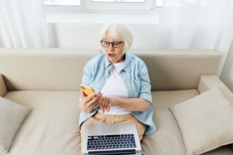 Senioren sitzt auf Sofa mit Handy und Computer und nutzt das Internet.