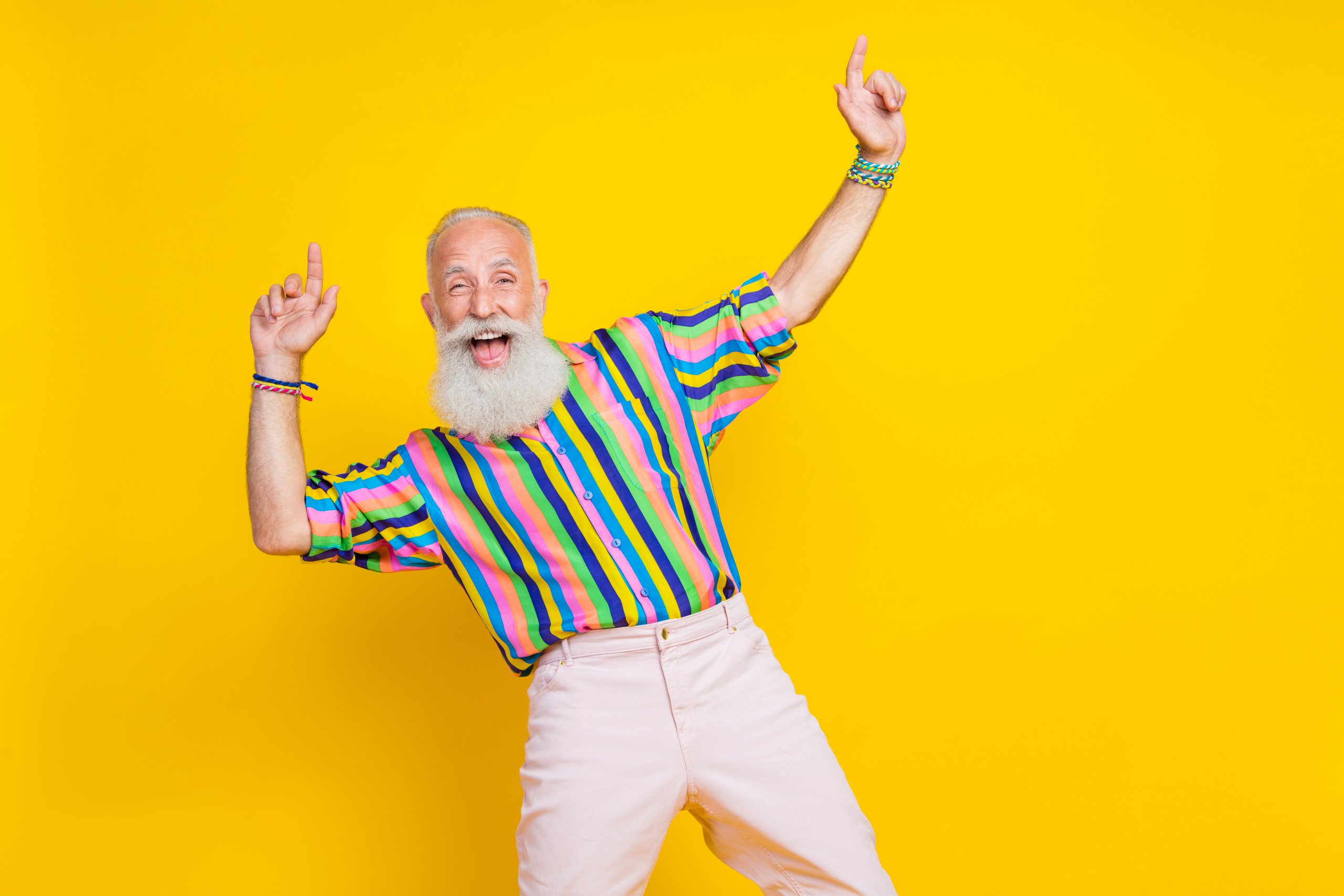 Ein bärtiger älterer Herr trägt ein buntgestreiftes Hemd, hebt die Hände zum Tanzrhythmus und lacht vor einem knallgelben Hintergrund.