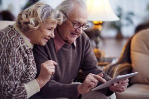 Eine ältere Dame und ein Herr mit Brille schauen auf ein Tablet. Der Mann tippt etwas ein und die Frau lächelt.