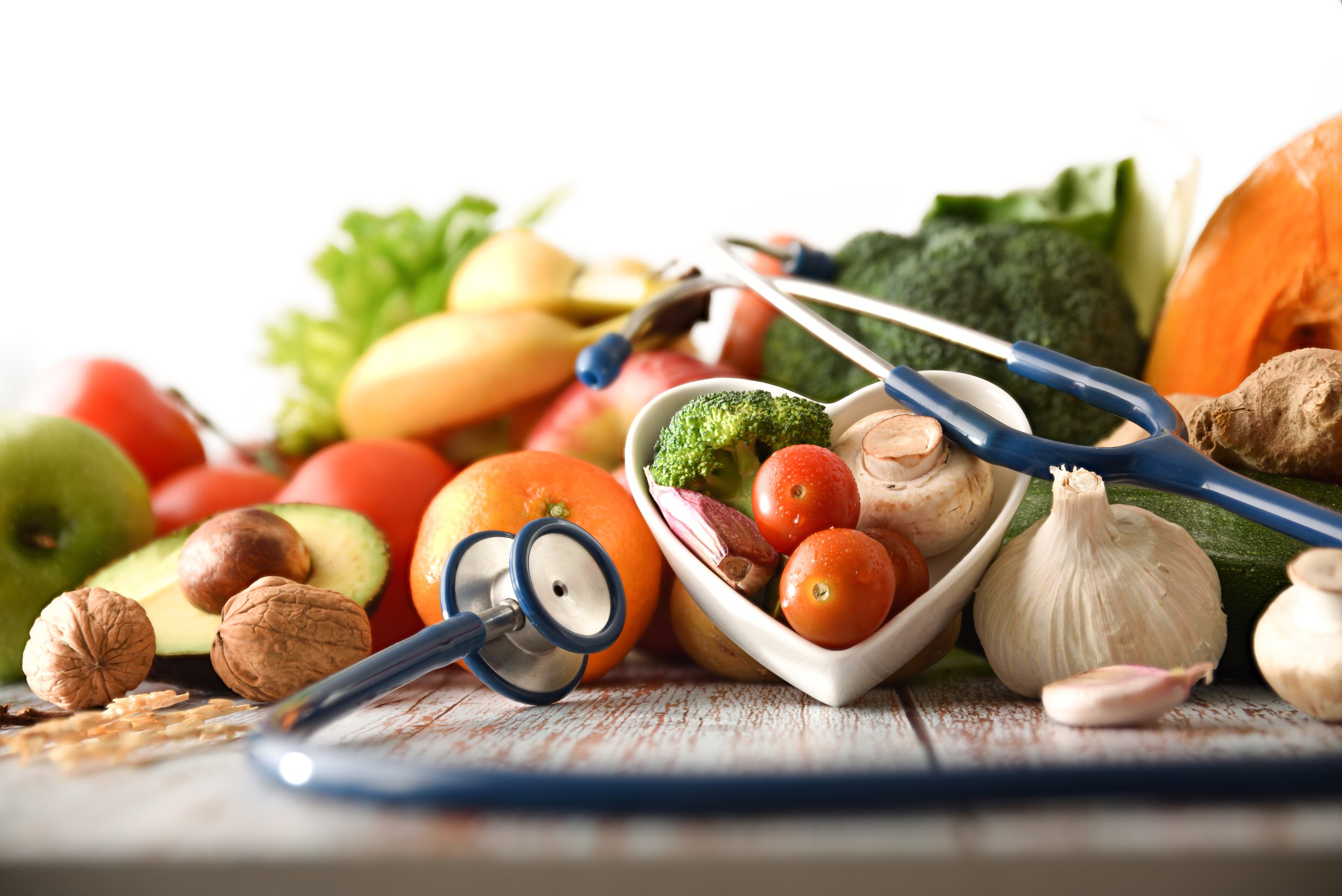 Eine bunte Gemüsemischung liegt auf einem Holztisch. In der Mitte ein Herz aus weißem Porzellan und ein Stethoskop.