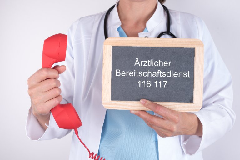 Ärztin in weißem Kittel hält rechts einen roten Telefonhörer in der Hand und links die Telefonnummer vom Bereitschaftsdienst.