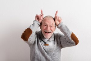 Älterer Herr in Strickpullover lacht und zeigt Hörner mit seinen Fingern.