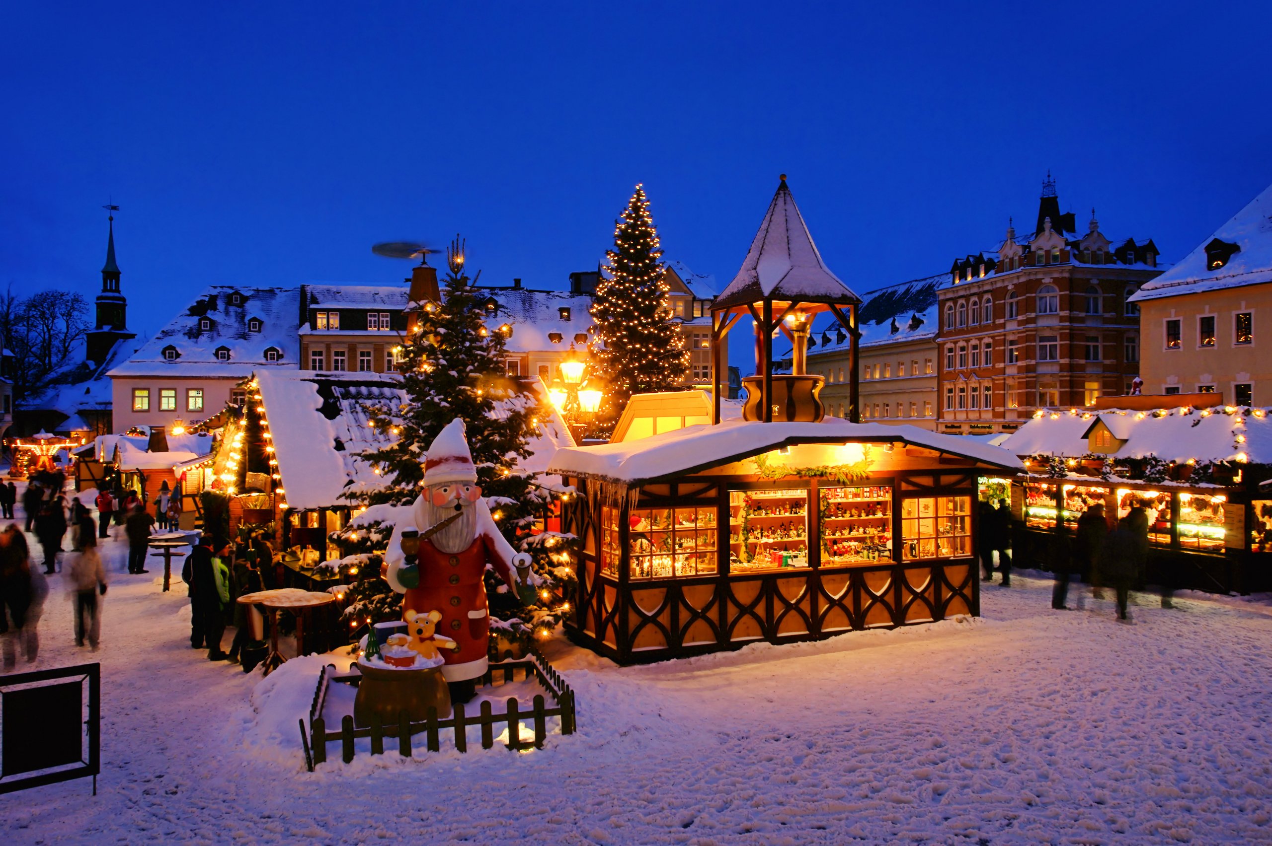 Festlich erleuchteter Weihnachtsmarkt, Schnee bedeckte Buden, geschmückte Tanne, riesiger Weihnachsmann mit Geschenkesack im Vordergrund. StockAdobe/LianeM