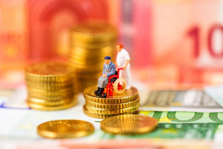 Ein Rollstuhlfahrer en miniature steht auf einem Turm aus Münzen und hinter ihm ein Pfleger in weißer Kleidung. Darunter 100 Euro-Scheine. Foto: AdobeStock, marcus_hofmann