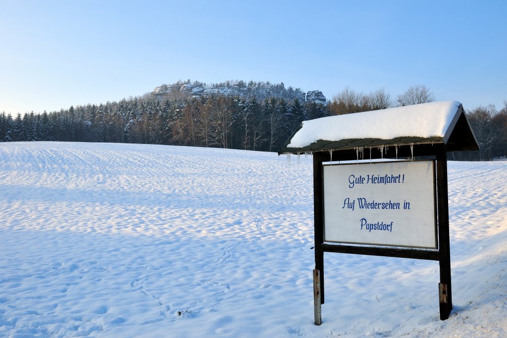 Schneebedecktes Feld, im Hintergrund Sandstein und Wald, rechts eine Tafel mit Gute Heimfahrt!