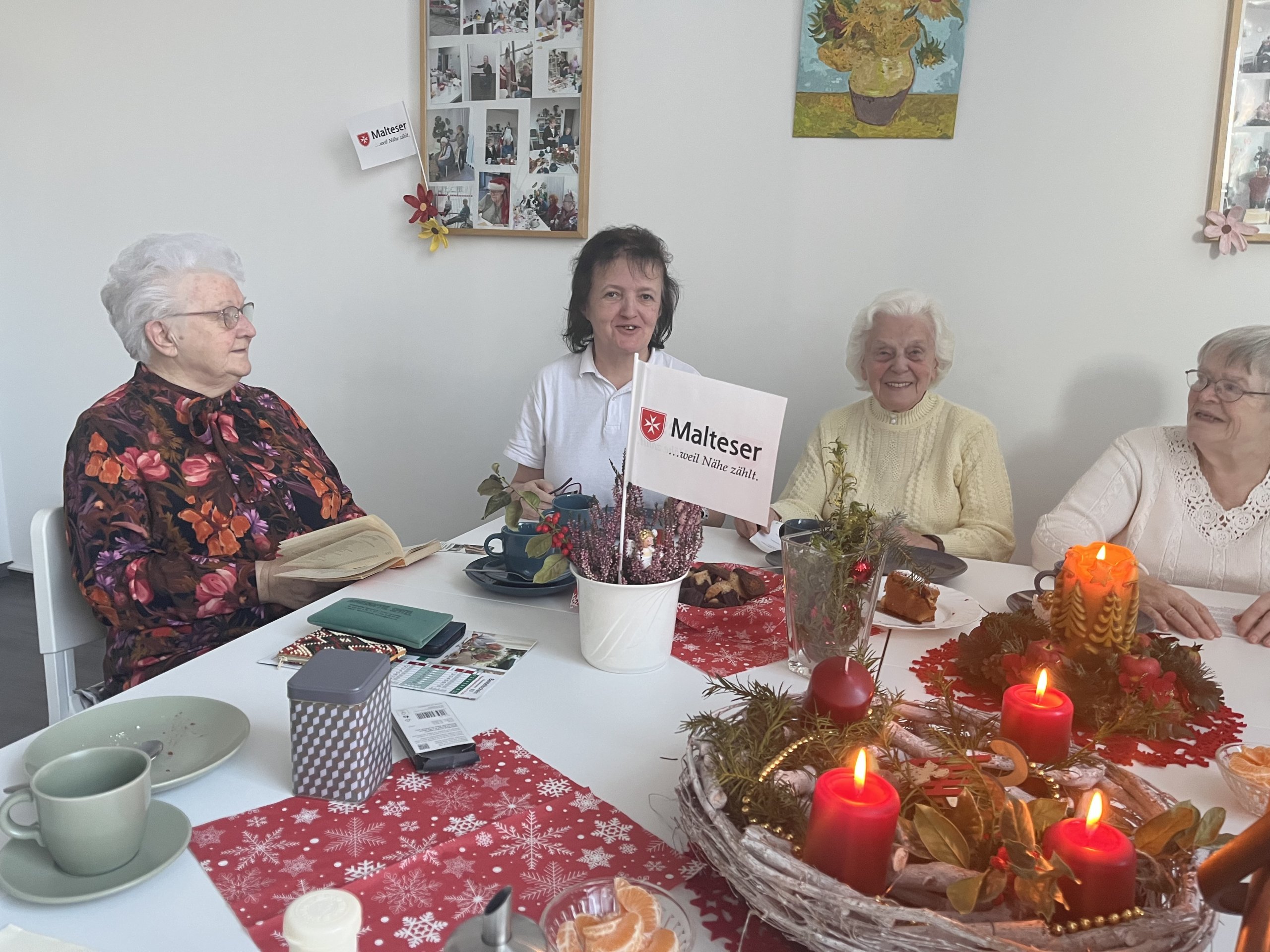 Eine Gruppe Seniorinnen und eine Betreuerin im weißen Kittel hat für das SZ-Lebensbegleiter-Foto Platz genommen. Im Vordergrund leuchten auf einem Adventskranz die roten Kerzen. Foto: Malteser
