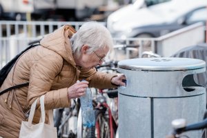 Eine ältere Dame sucht im Papierkorb einer Stadt nach leeren Pfandflaschen. Sie trägt eine beigefarbene Jacke und hat mehrere Tüten am Arm hängen.