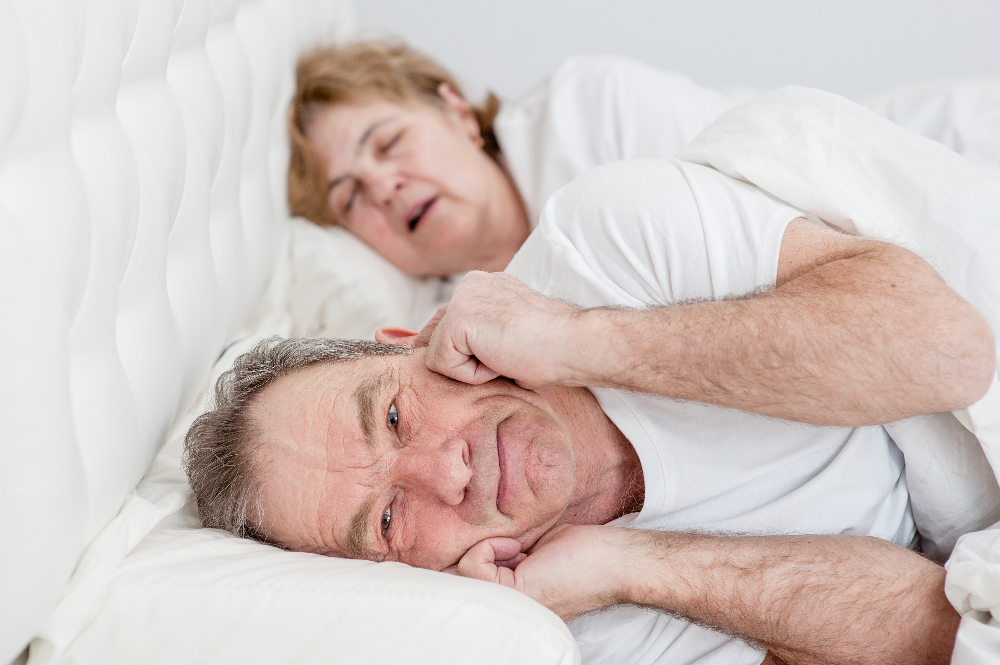 Älteres Paar liegt im Bett. Sie schläft mit offenem Mund und er ist wach und hält sich die Ohren zu.