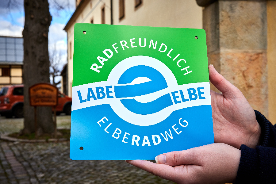 Die Plakette "Radfreundlich Elberadweg" in grün und blau zeigt den Hinweis für radfreundliche Unterkünfte auf der Strecke am Elberadweg.