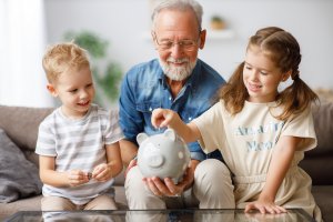 Ein Großvater hilft seinen Enkeln Geld sparen. Münzen werden in ein Sparschwein geworfen. Foto: AdobeStock/JenkoAtaman