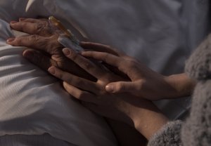 Frau hält mit beiden Händen die Hände einer hochaltrigen Person auf der Bettdecke.