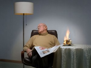 Ein Senior hält eine Zeitung in der Hand und ist eingeschlafen. Das Papier hat an einer Kerze Feuer gefangen. Foto: Forum Brandrauchprävention