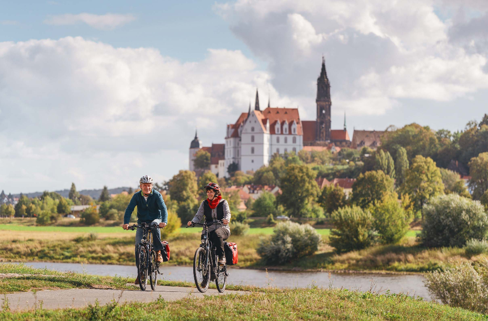 Ein älteres Paar ist mit einem Fahrrad mit Satteltaschen unterwegs auf dem Elberadweg. Im Hintergrund ist die Albrechtsburg in Meißen vor einem leicht bewölktem blau-weißen Himmel zu sehen.
