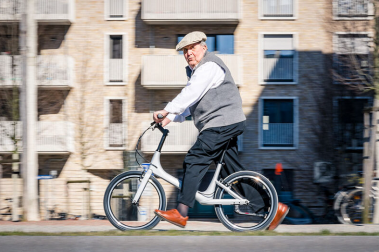Ein älterer Herr mit Pullunder und Schirmmütze läuft mit einem Laufrad durch die Stadt.